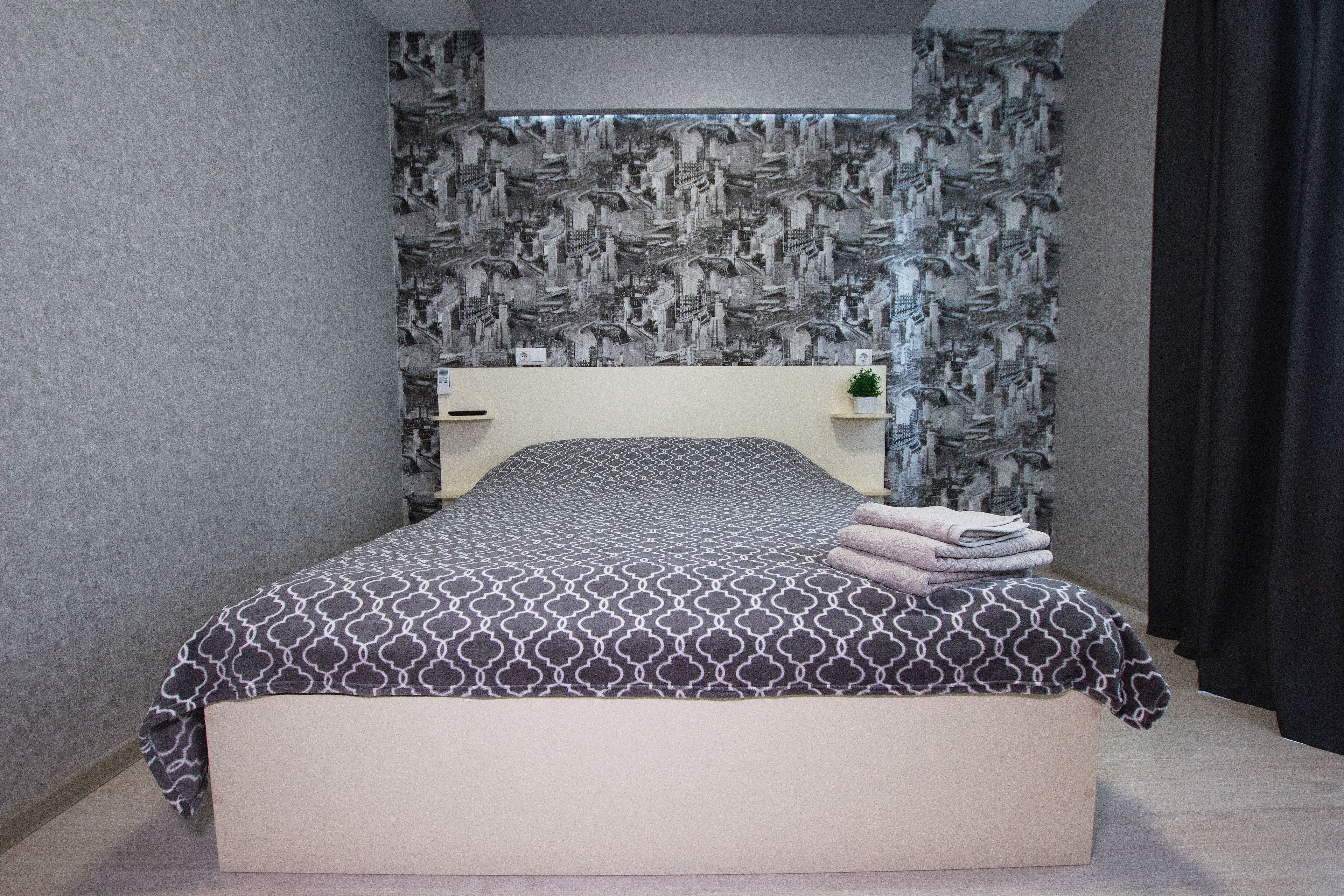 A neatly made bed with a patterned bedspread, a headboard, and a small shelf with a plant.