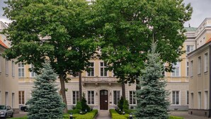 Un edificio con facciata simmetrica, ingresso centrale e un giardino curato con siepi potate e due grandi alberi sempreverdi.
