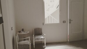 Ein Zimmer mit weißer Tür, einem kleinen Tisch mit Stuhl und einem Fenster mit Blick auf eine Treppe.