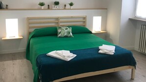 Ein ordentlich bezogenes Bett mit grüner Bettwäsche, zwei Nachttischlampen, ein Regal mit kleinen Pflanzen und einem 'LOVE'-Schild, und ein Fenster auf der rechten Seite.
