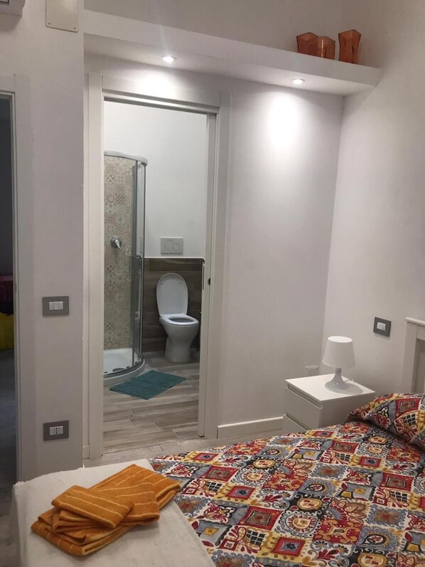 A bedroom with a bed, a bedside table with a lamp, a bathroom with a shower and toilet, and a door leading to another room. - Appartamento Badia (Foligno)