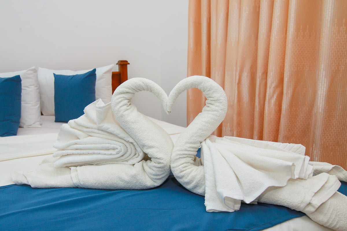 A bed with white towels arranged in a swan shape, blue pillows, and an orange curtain.