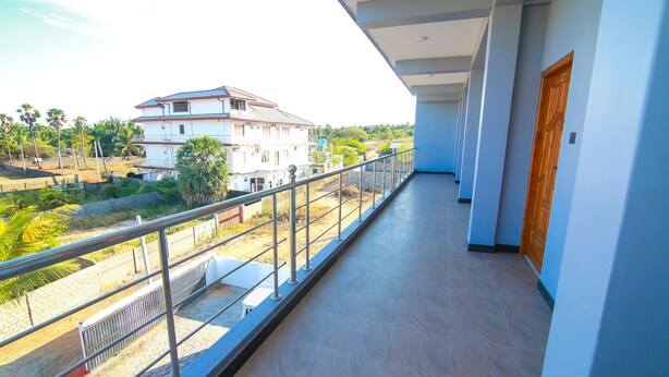 A balcony with a metal railing, a view of a residential area with houses and greenery, and a door on the right side. - Hotel Sun Blue (Trincomalee)