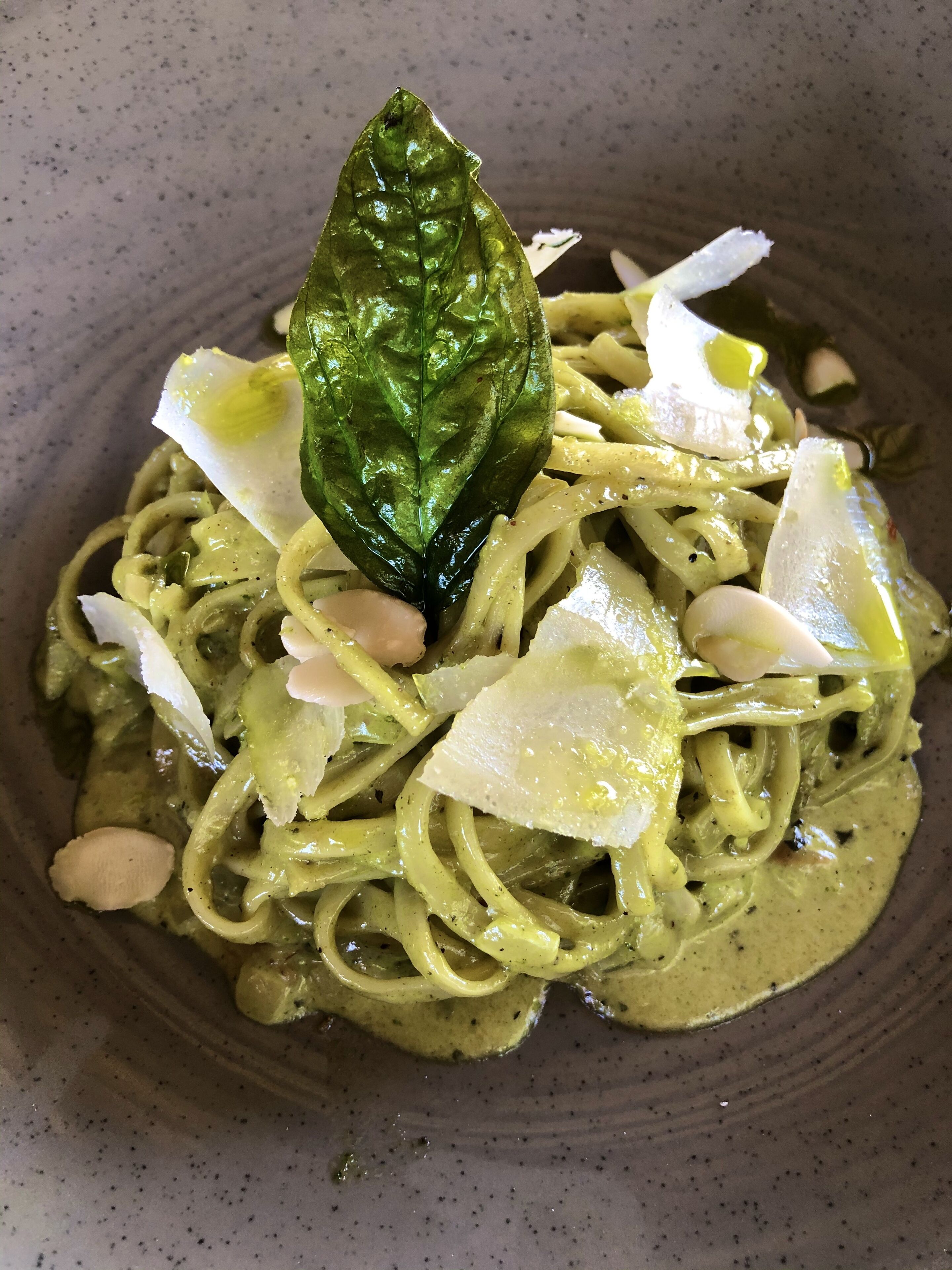 A bowl of pasta with a green sauce, garnished with a basil leaf and shaved cheese.