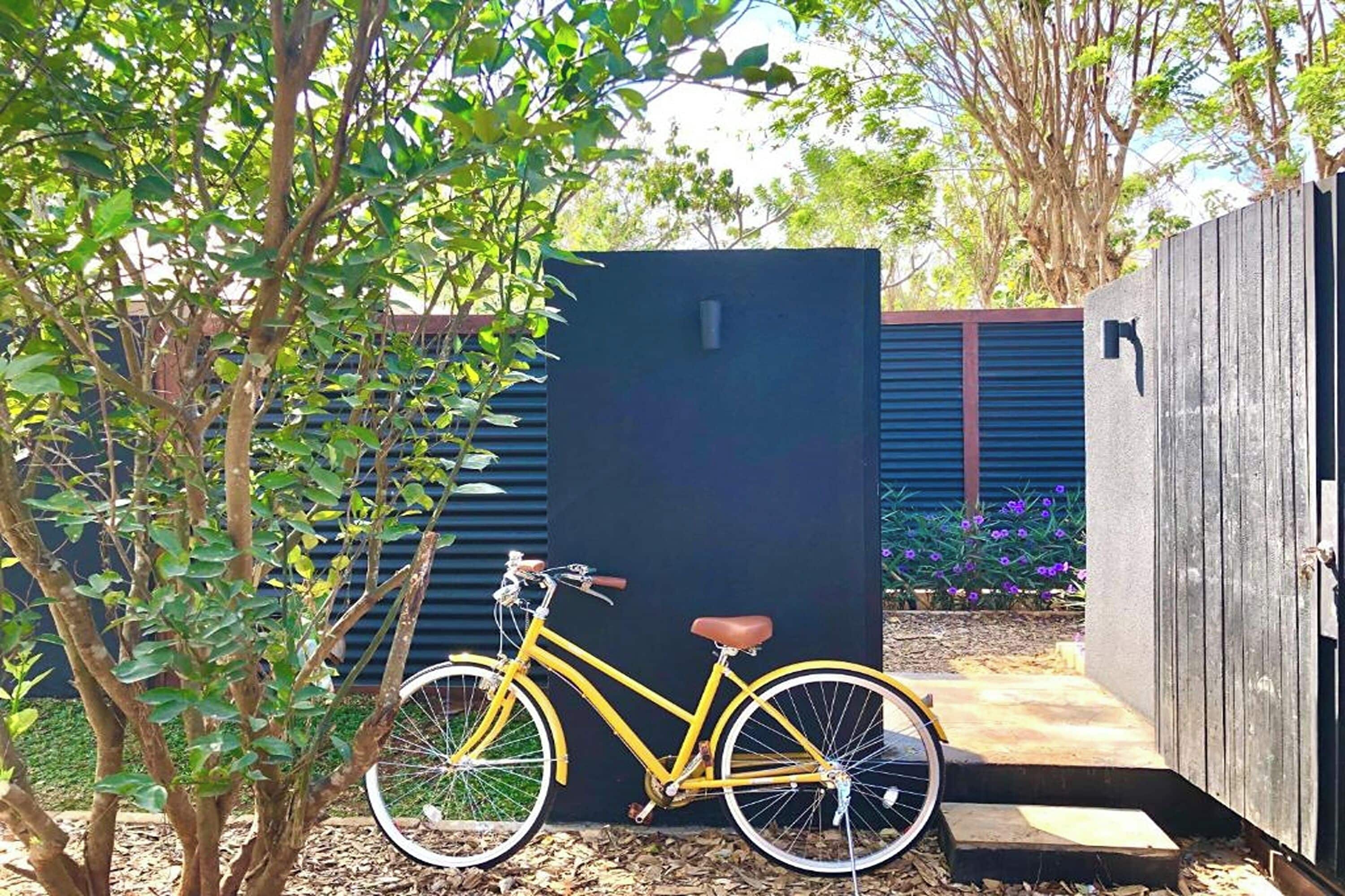 A yellow bicycle parked next to a black structure, surrounded by greenery and a wooden fence.