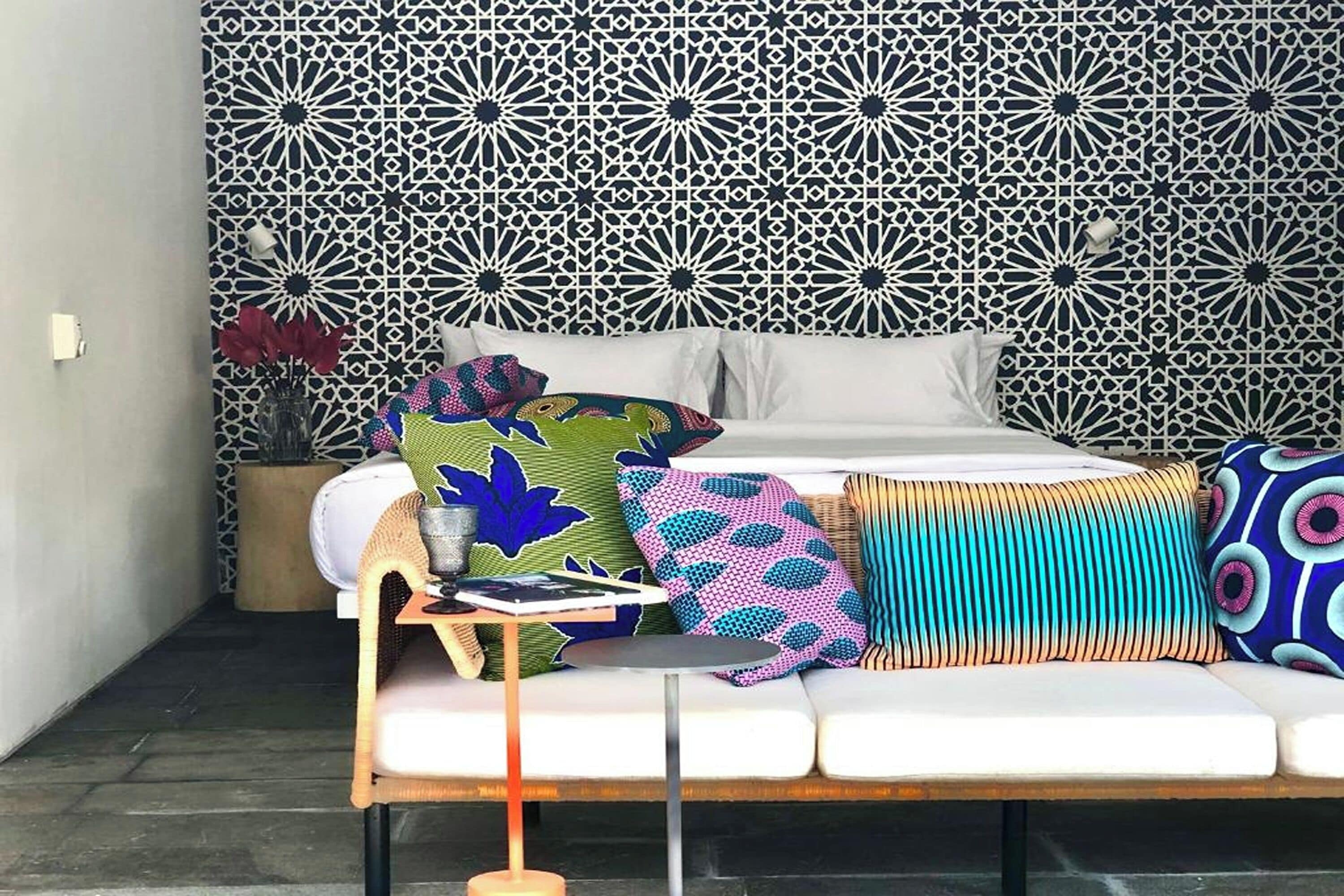 A modern room with a patterned wall, a sofa with colorful cushions, a bedside table with a book and a glass, and a potted plant.