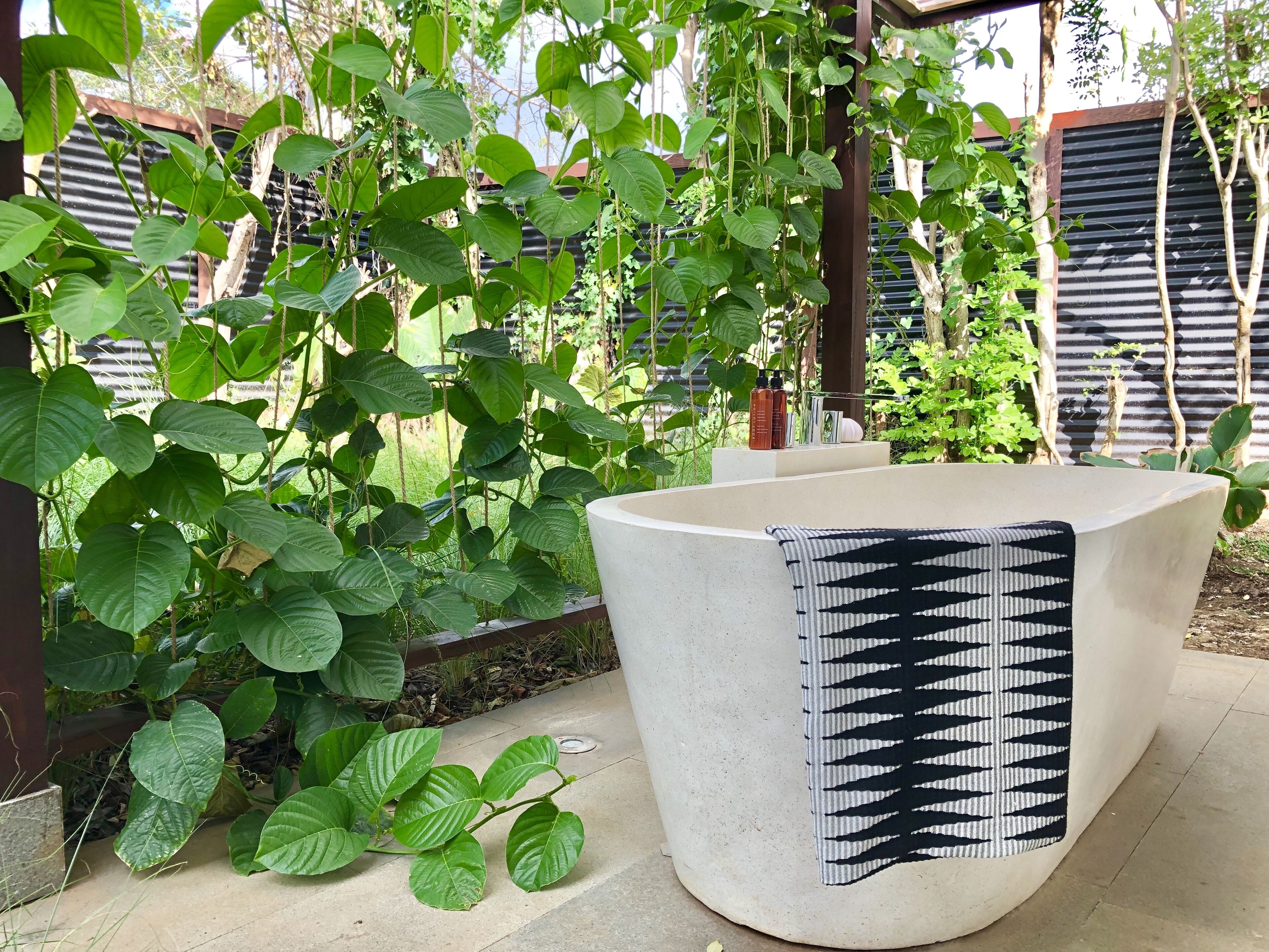 A concrete planter with a patterned cushion, surrounded by lush green plants and a metal fence.