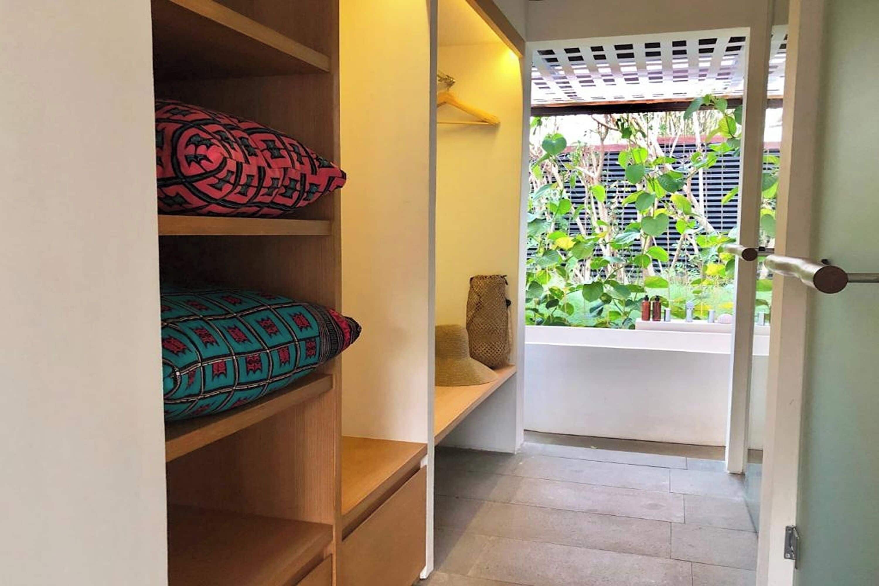 A room with a wooden shelf with patterned cushions, a bench, and a view of greenery through a window.