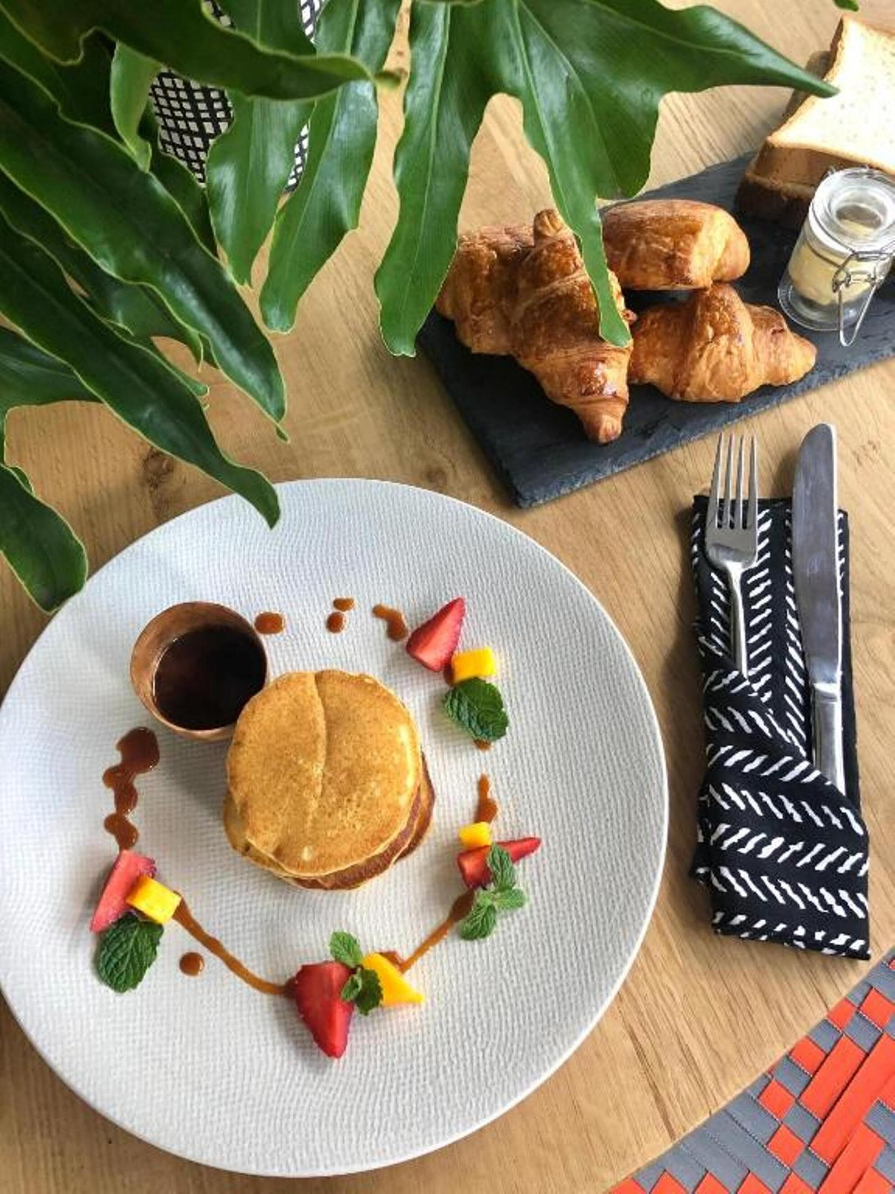 A breakfast setting with pancakes, strawberries, and mint leaves on a plate, accompanied by croissants and a small jar of jam on a napkin.