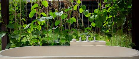 A large, empty bathtub in a garden setting with lush greenery and a metal fence in the background.