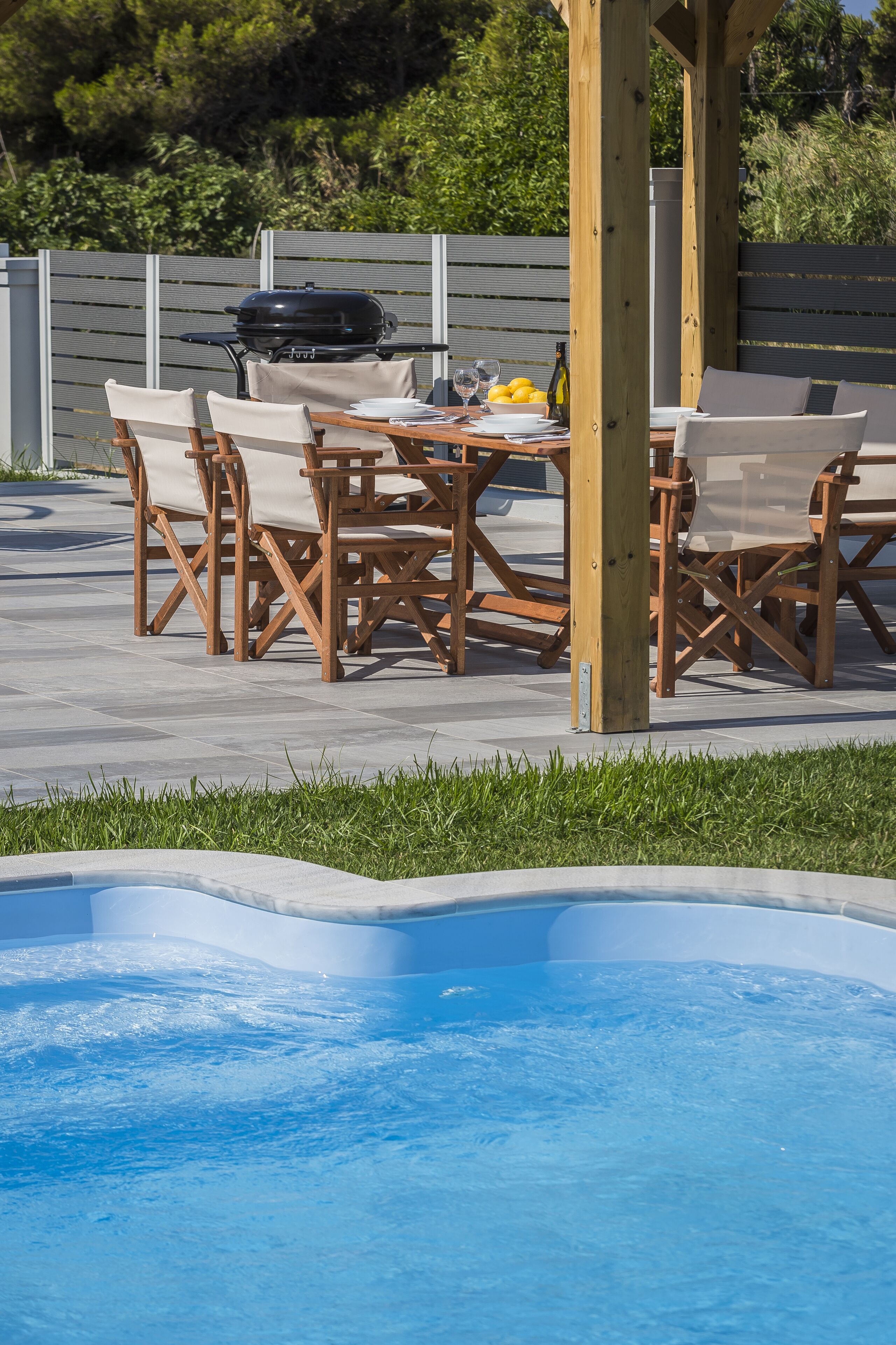 Une terrasse extérieure avec une piscine, des chaises en bois, une table et un barbecue.