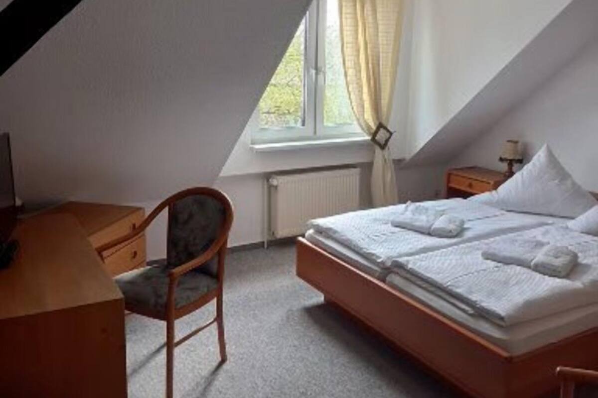 Un dormitorio con techo inclinado, una cama con almohadas y toallas, un escritorio de madera, una silla y un ventanal con cortinas.