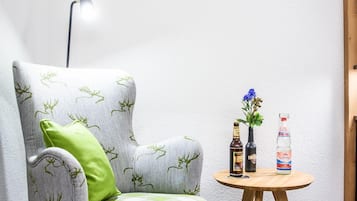 A corner room with a grey armchair with green cushions, a small wooden table with two bottles and a vase, and a floor lamp.