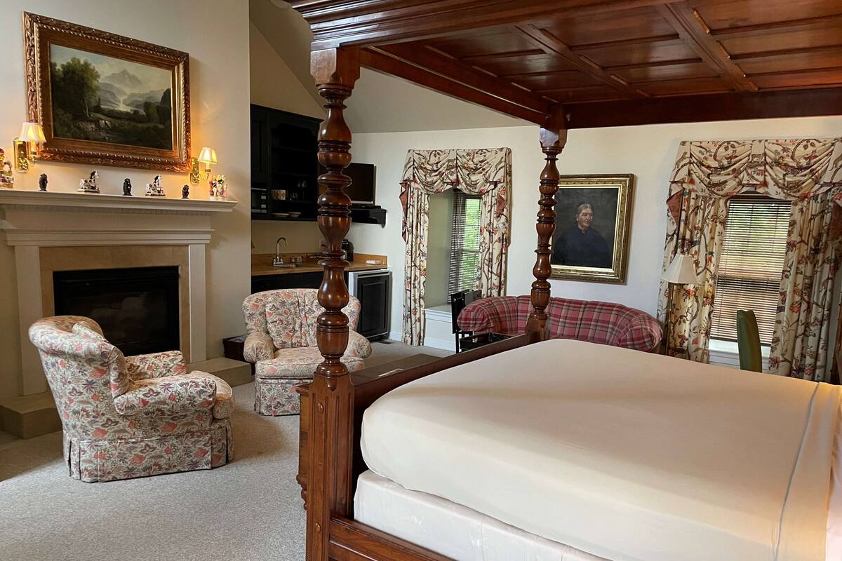 Une chambre à coucher avec un lit à baldaquin, une cheminée, des fauteuils à motifs floraux et un tableau accroché au mur.