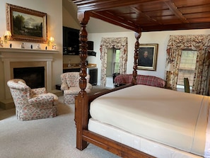 A bedroom with a four-poster bed, a fireplace, floral armchairs, and a painting on the wall.