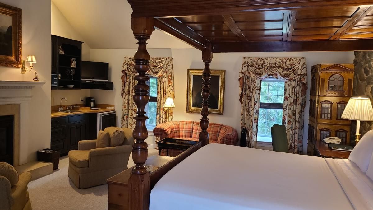 A hotel room with a four-poster bed, a sofa, a fireplace, a kitchen area, and a view of the outside.
