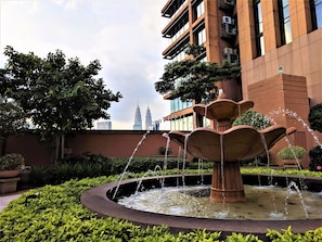 Surrounded by lush greenery, this tranquil outdoor space features a central water fountain and a striking modern building with a prominent tower.