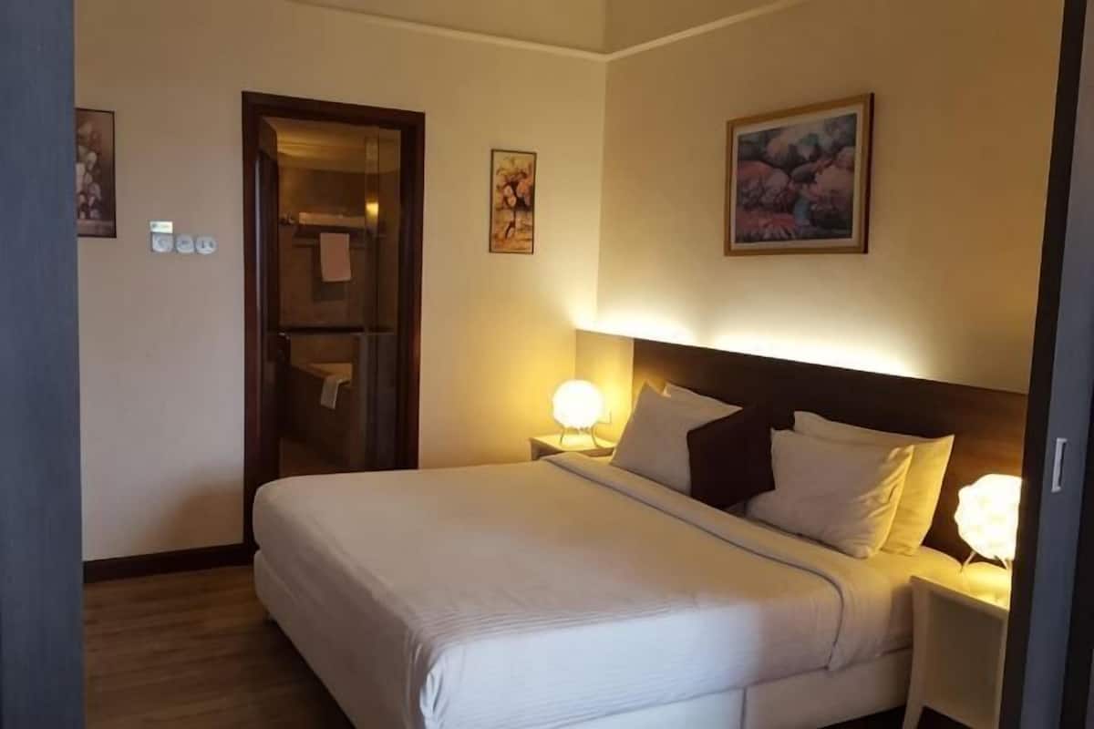 A hotel room with a bed, bedside lamps, a door leading to a bathroom, and a painting on the wall.