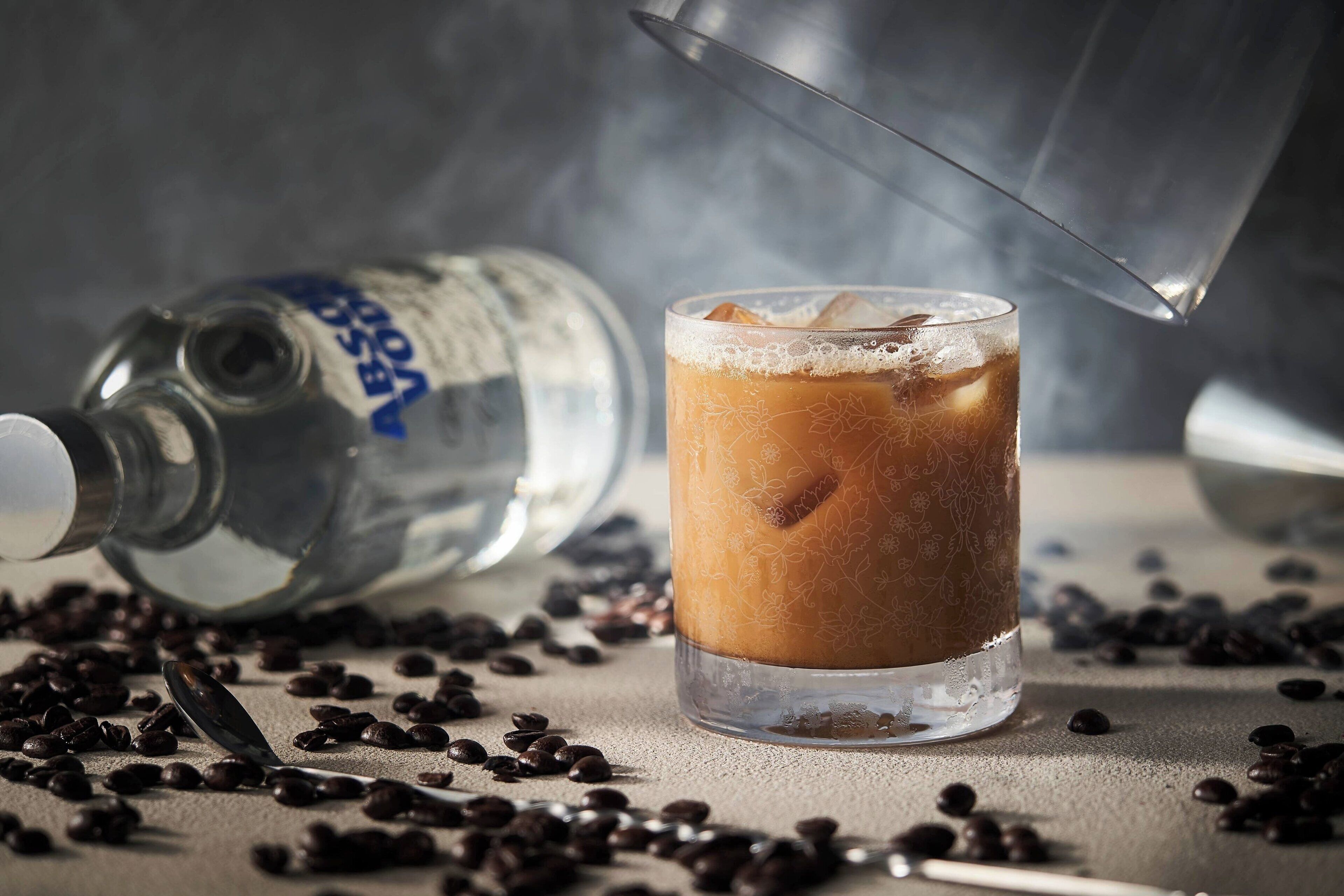 Un vaso de café helado con una jarra de metal volcadas, rodeado de granos de café sobre una superficie rugosa.