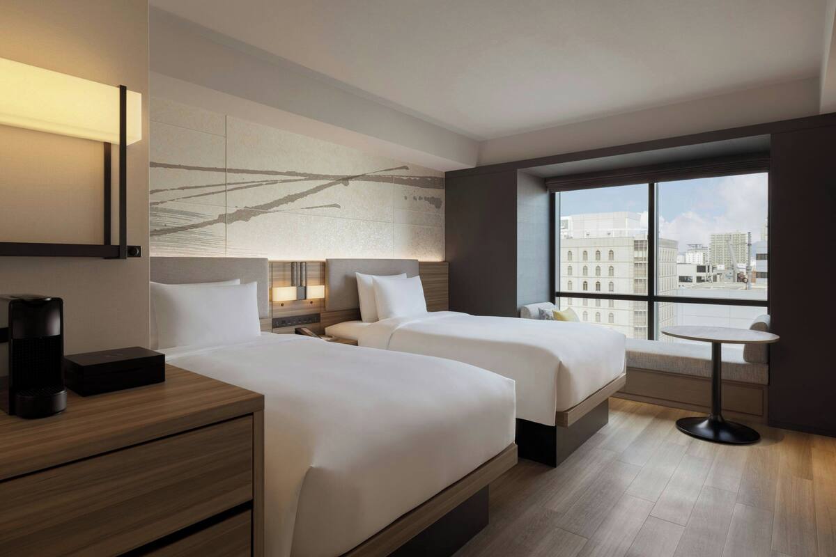 A modern hotel room with two beds, a bedside table with a lamp, a small round table, and a large window offering a city view.