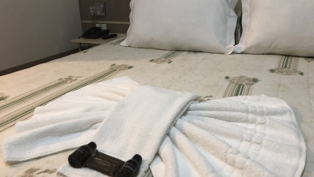 En pænt redt seng med hvide sengetøj, et par sorte slippers og en lotionflaske på sengen.