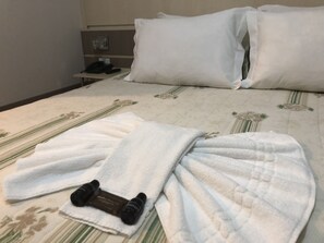 A neatly made bed with white linens, a pair of black slippers, and a bottle of lotion on the bed. - Elite Palace Hotel (Prudentopolis)