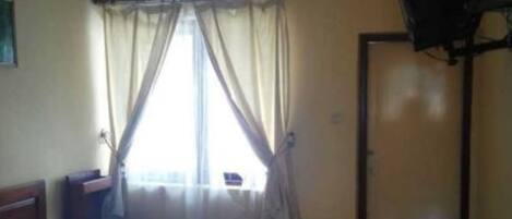 A room with a bed, a television on the wall, a window with curtains, and a door.