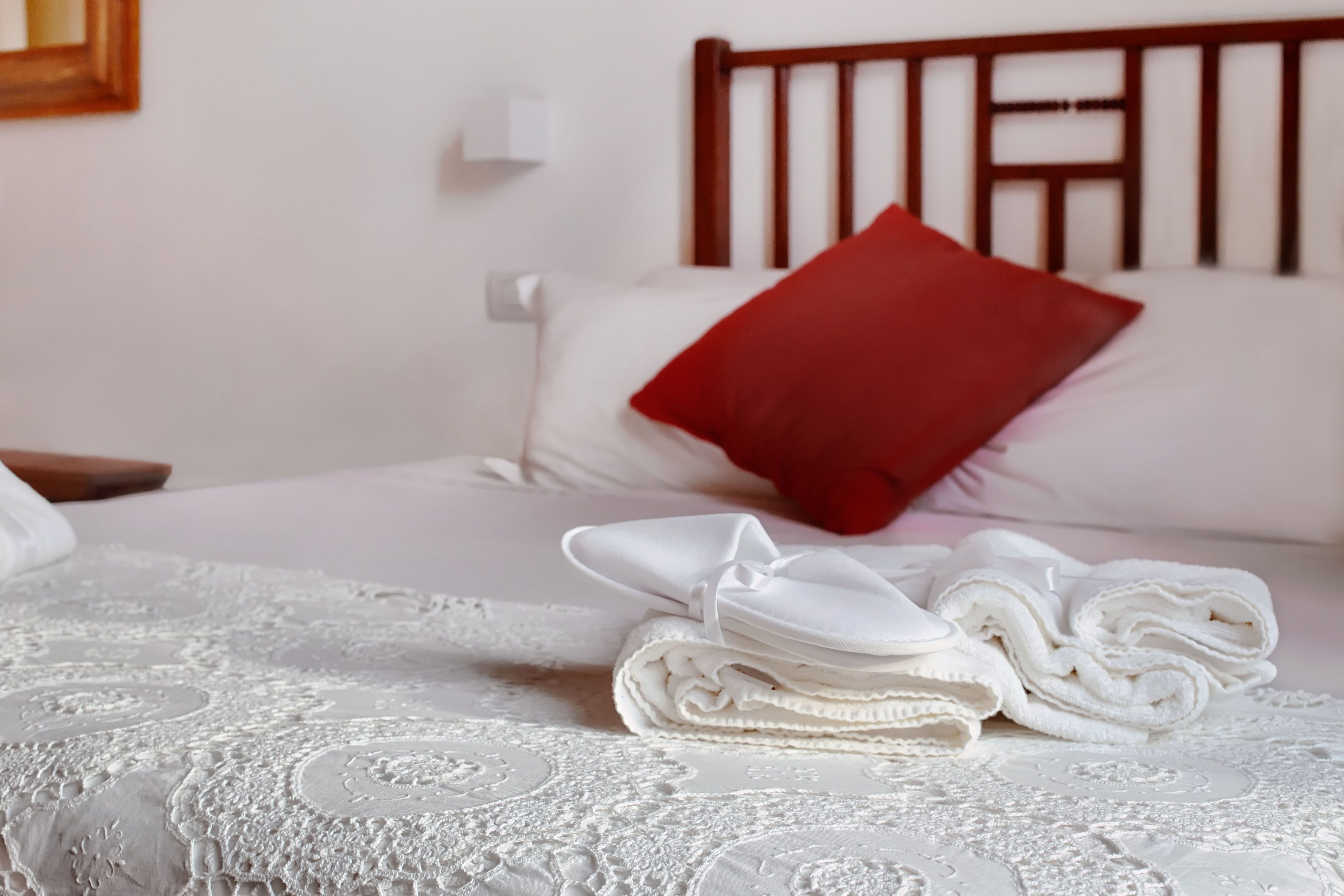 A neatly made bed with a white bedspread and a red pillow, a folded white towel, and a white pillow on a wooden headboard.
