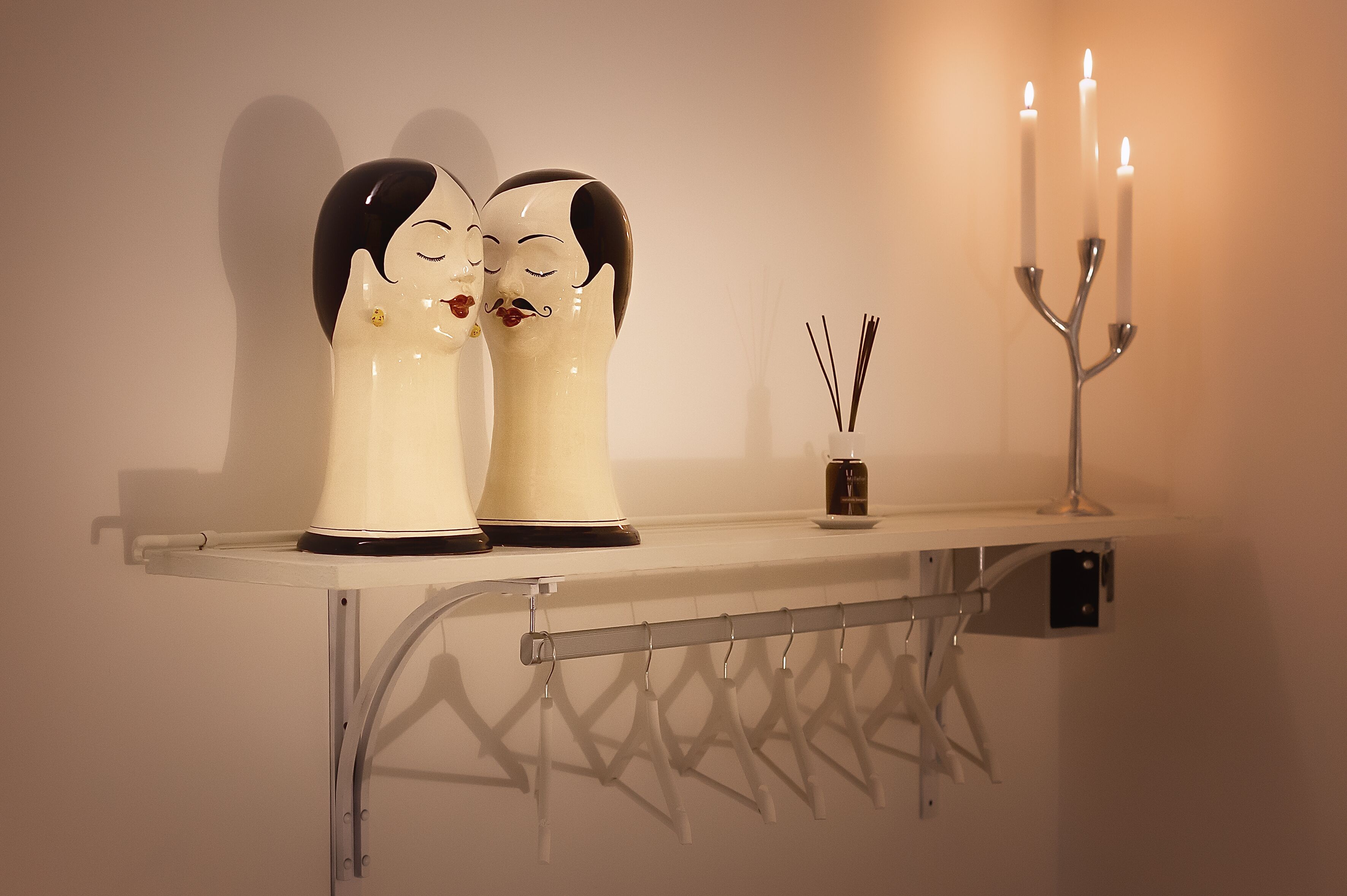 Uma prateleira com duas figuras de cerâmica, um castiçal com três velas e um pequeno recipiente com varetas de junco.