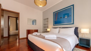 Una cama bien hecha con sábanas blancas y un edredón color beige, una mesita de noche con lámpara, un cuadro abstracto enmarcado en la pared y suelo de madera.