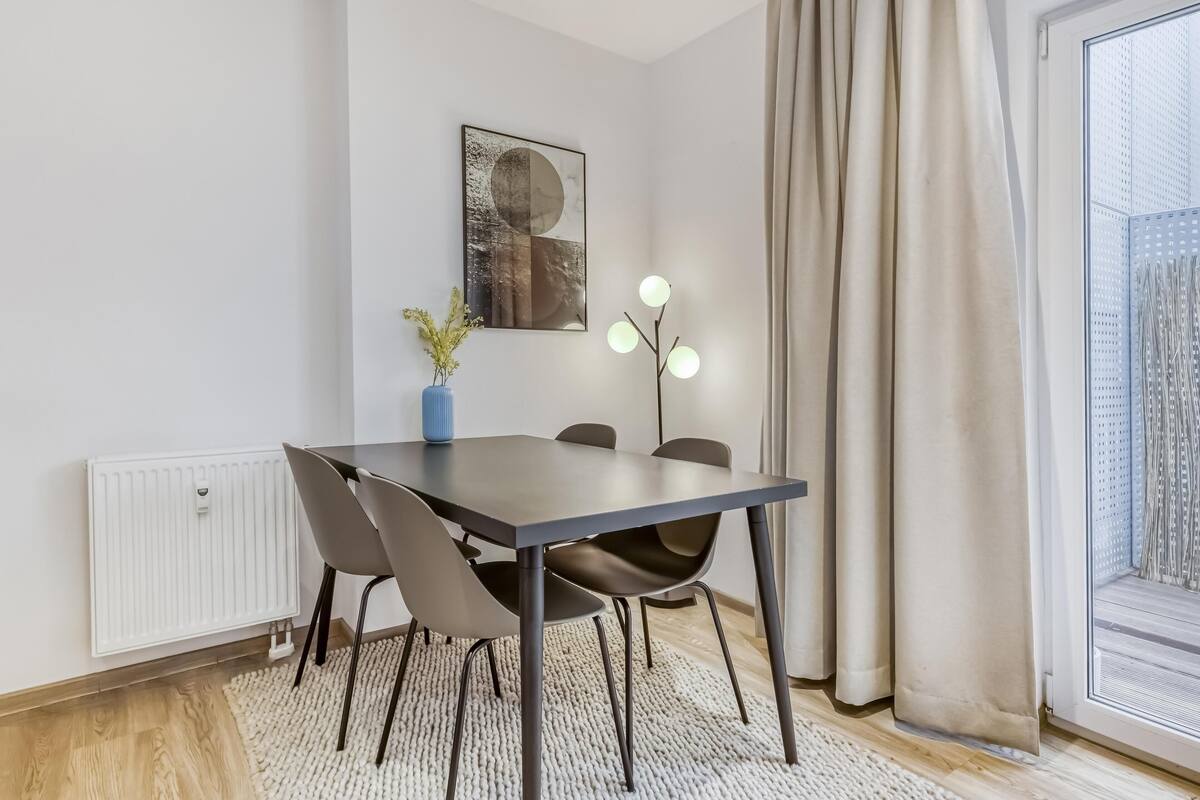 Ein moderner Essbereich mit einem dunklen Tisch, vier Stühlen, einer blauen Vase mit Pflanzen, einer Wandleuchte und einem Fenster mit Vorhängen.