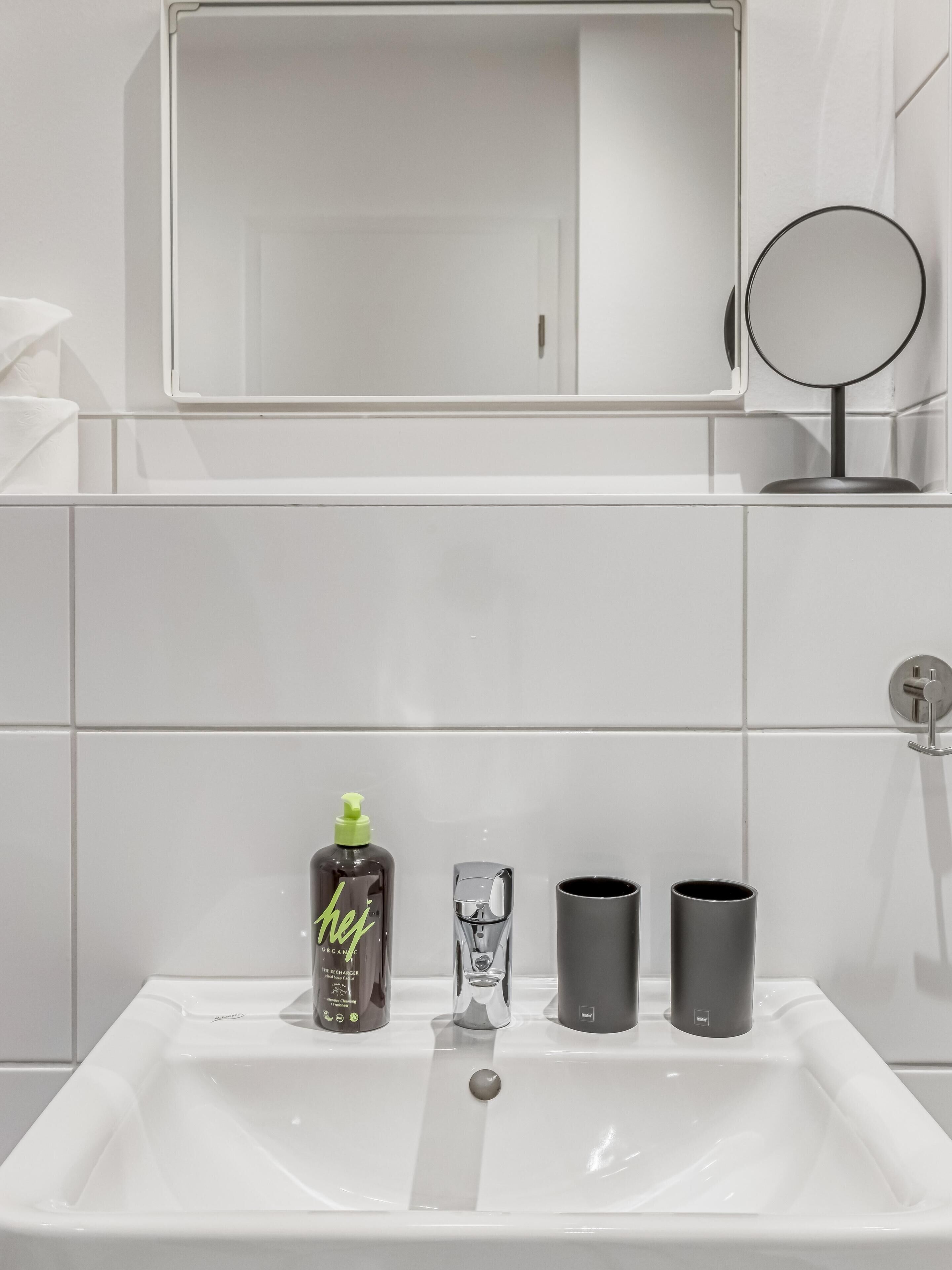 Een badkamer met een wit wastafel, een spiegel en toiletartikelen, waaronder een fles, een glas en twee bekers.
