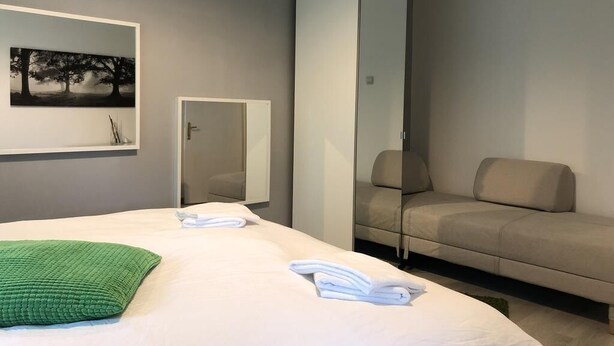 Ein modernes Hotelzimmer mit einem Bett, einem grünen Kopfkissen, einer beigen Schlafsofa, einem großen Spiegel und einem gerahmten Bild an der Wand.