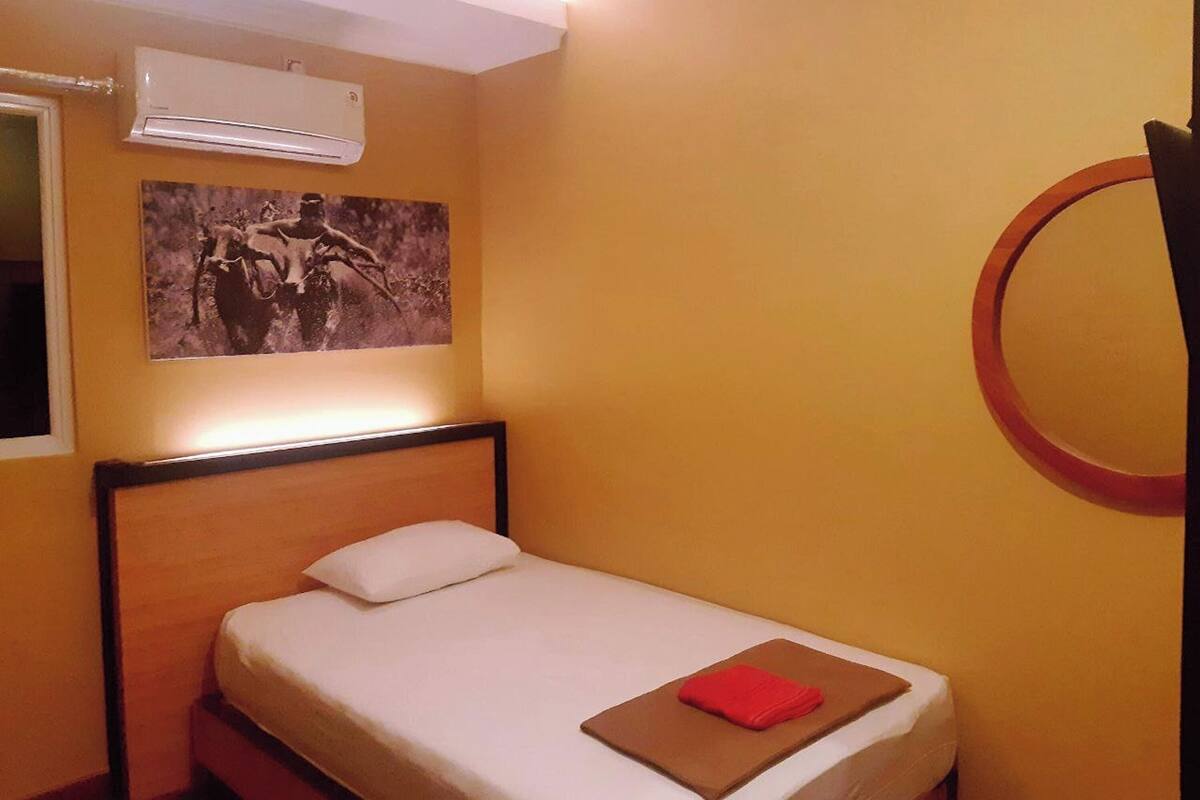 A single bed with a white mattress, a red towel, and a framed picture on the wall.