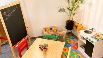 Un cuarto de juegos infantil con una mesa de madera, sillas, una pizarra, un juego de cocina para niños y una planta en maceta.