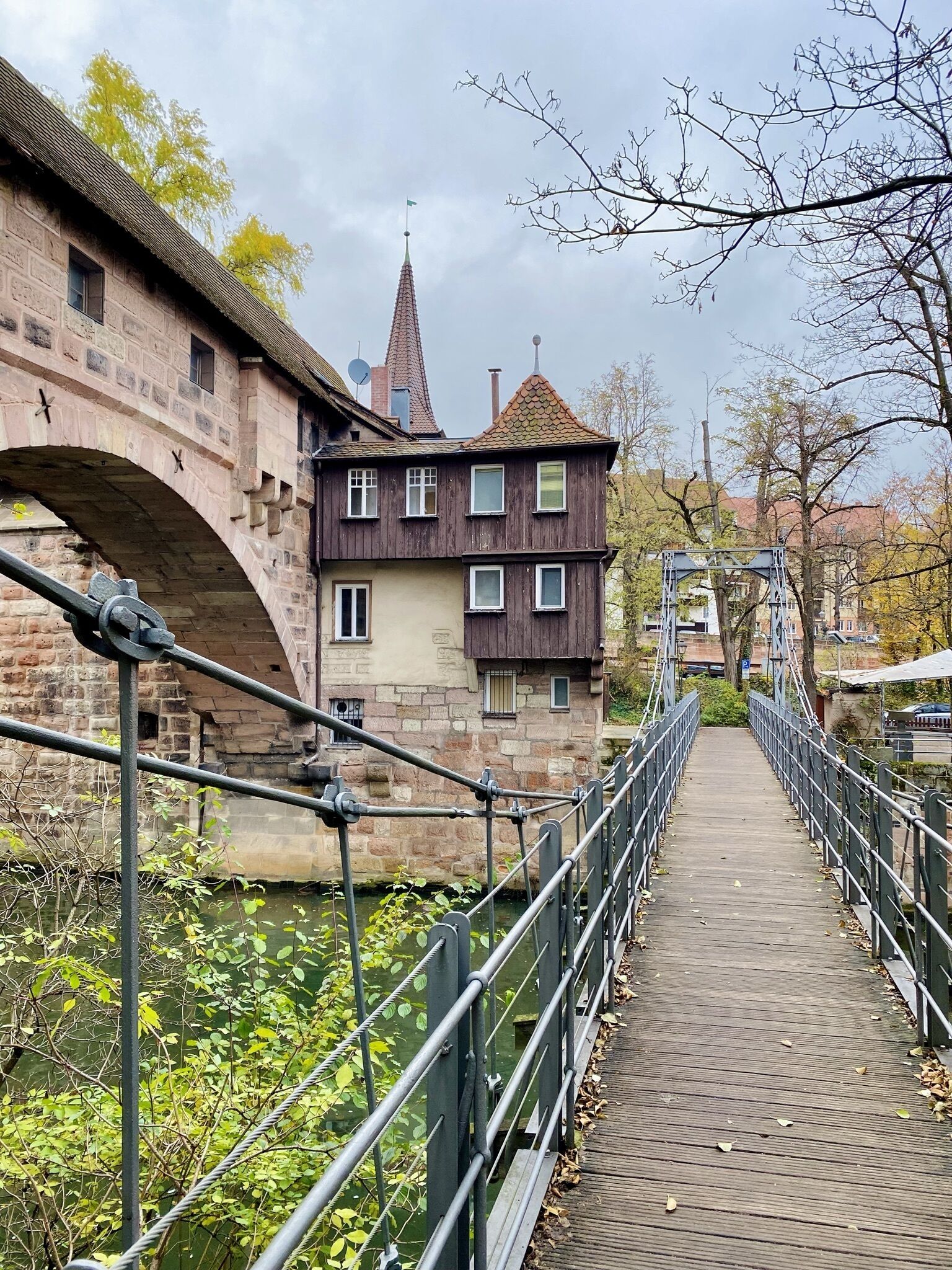 Un puente de madera con barandillas metálicas que cruza una masa de agua, conduciendo a un edificio con una torre y múltiples ventanas.