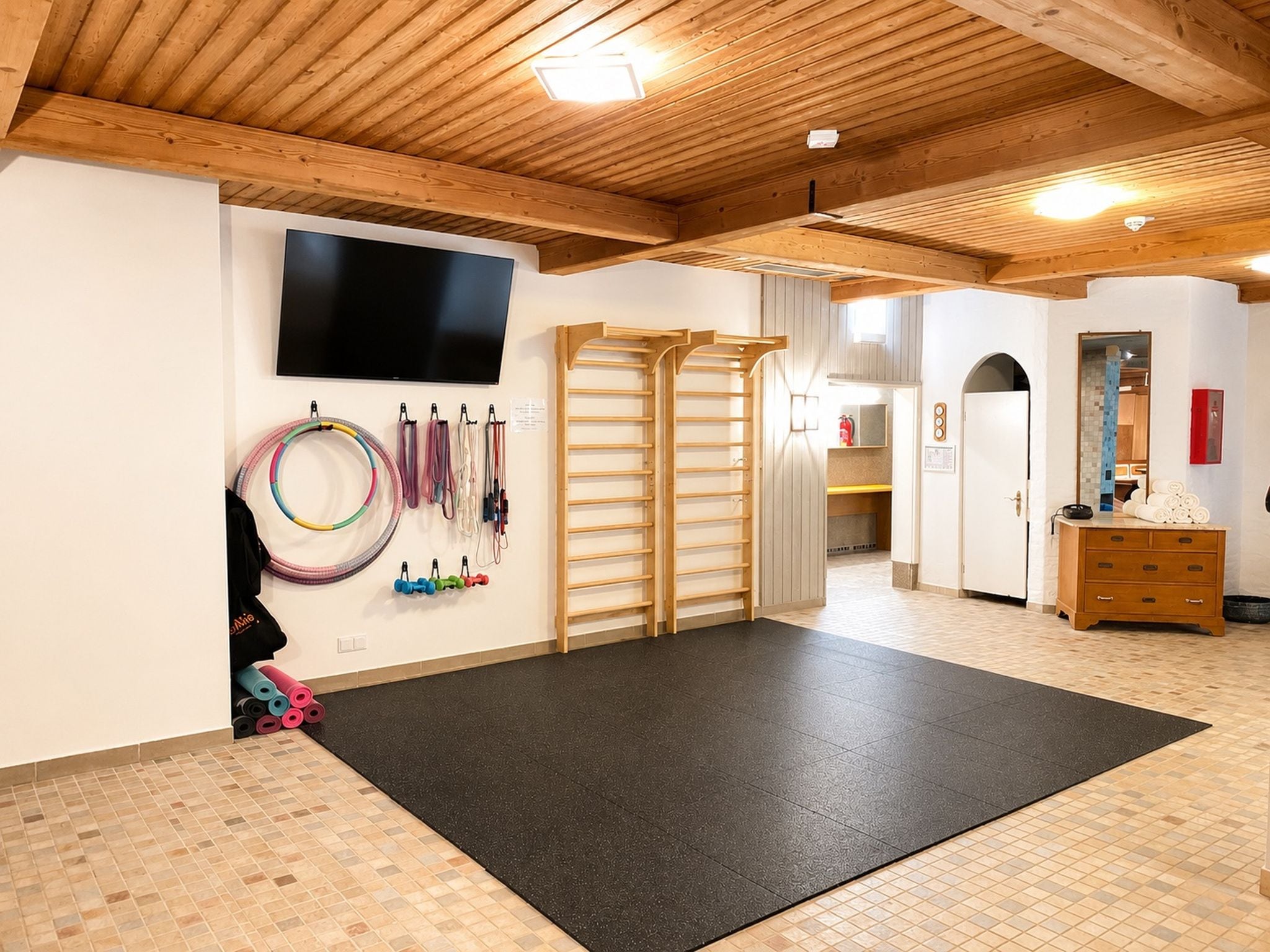 Ein Fitnessraum mit Trainingsgeräten, einer schwarzen Yogamatte und einem Fernseher an der Wand.