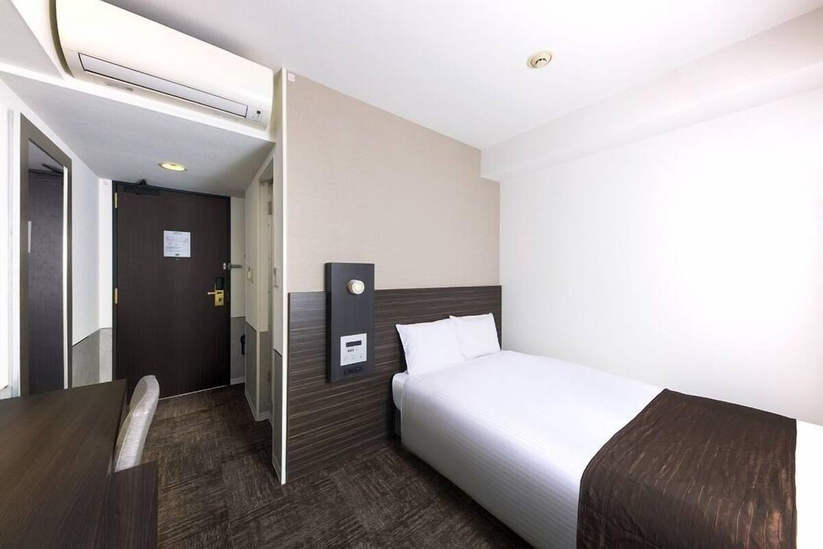 Une chambre d’hôtel équipée d’un lit, d’un bureau, d’une chaise, d’une porte et d’un panneau de commande fixé au mur.