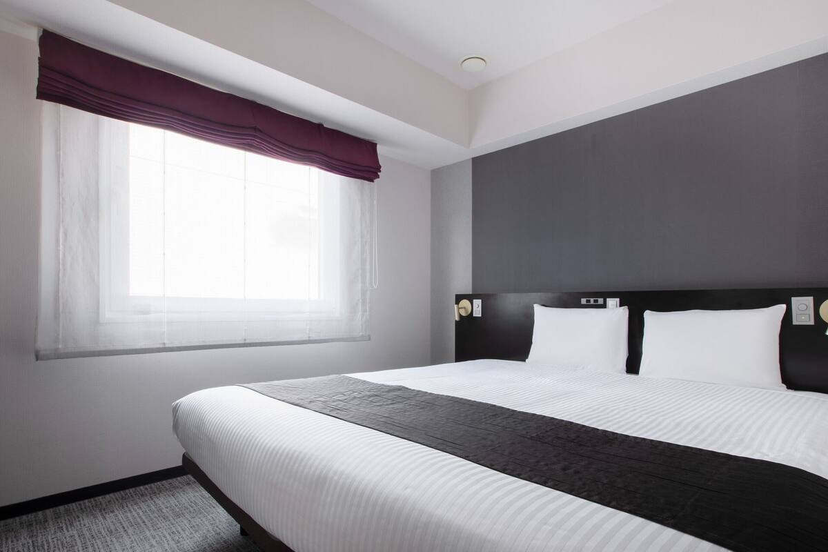Une chambre d’hôtel avec un grand lit, une fenêtre ornée de rideaux légers, une tête de lit sombre et une décoration aux couleurs neutres.