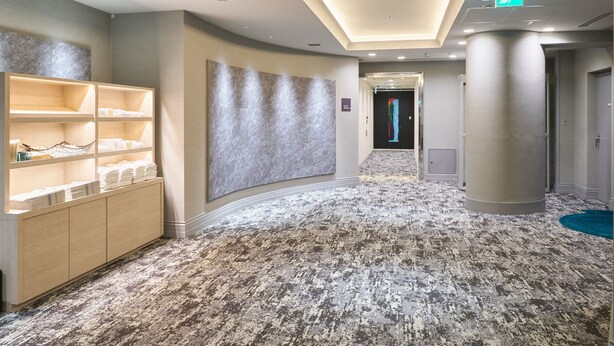 A hotel corridor with a textured carpet, a reception area with shelves, a wall-mounted artwork, and an emergency exit sign. - Hotel Villa Fontaine Tokyo - Nihombashi Mitsukoshimae (Tokyo)