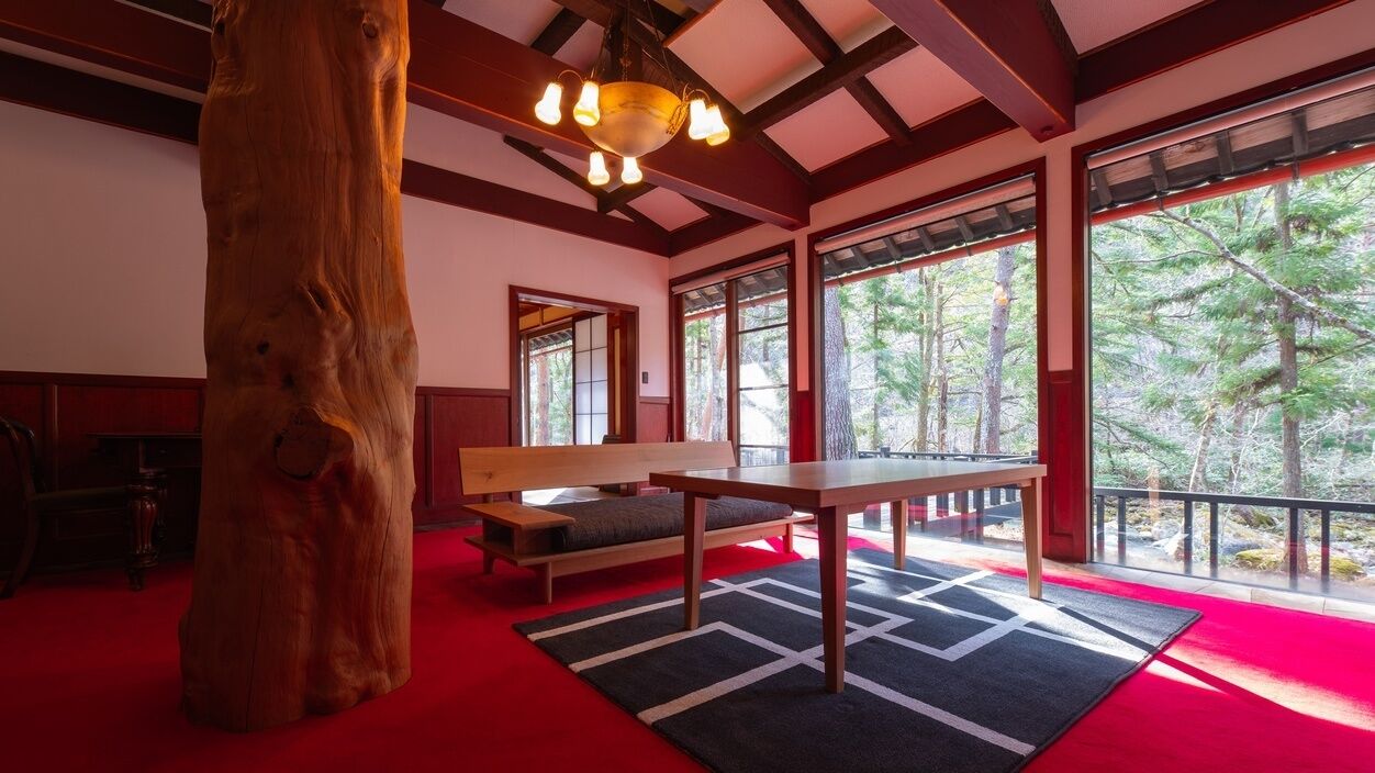 Una habitación con un gran tronco de árbol, una banca, una mesa y amplias ventanas con vistas a árboles.