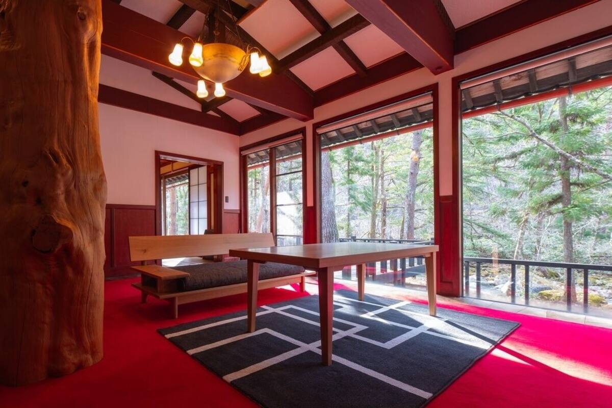 A room with a large wooden tree trunk, a bench, a table, and large windows with a view of trees.