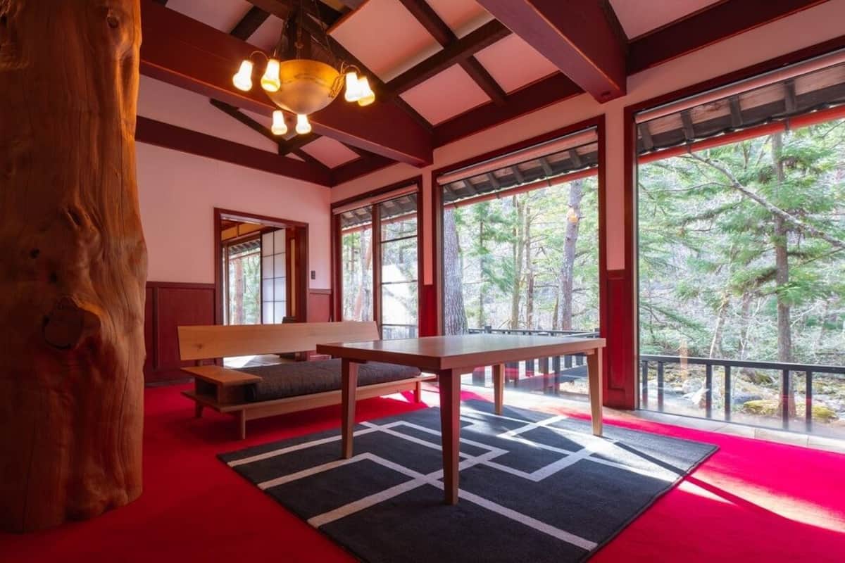 A room with a large wooden tree trunk, a bench, a table, and large windows with a view of trees.