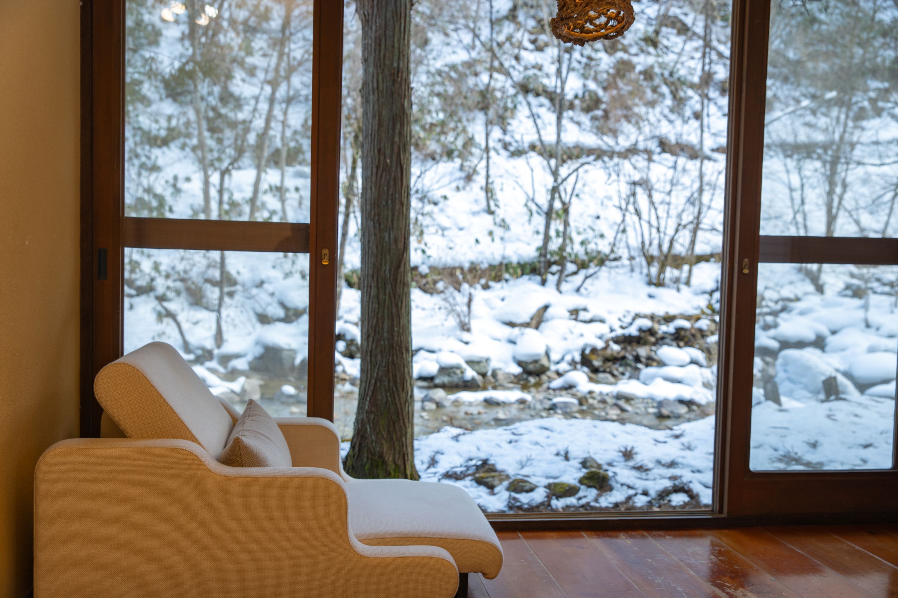 A room with a large window overlooking a snowy landscape, a wooden floor, and a beige sofa with a pillow.