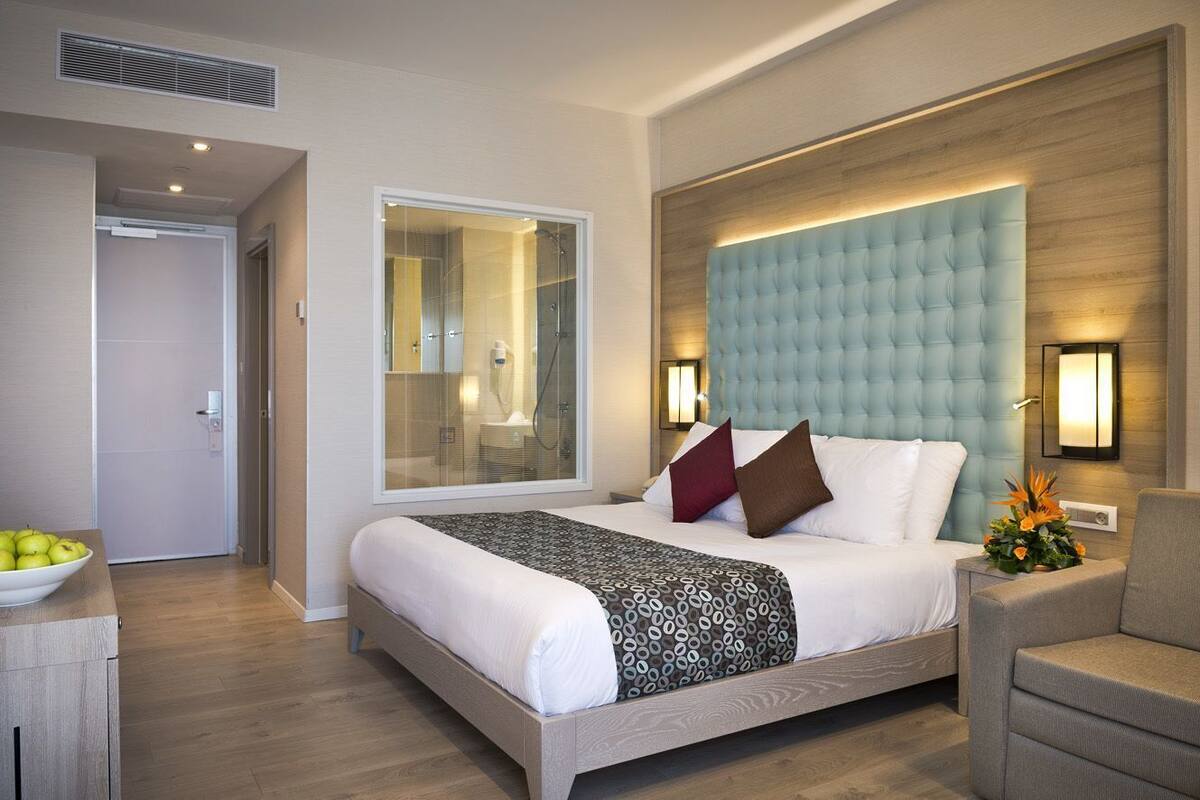 Una habitació d'hotel moderna amb un llit gran, una zona de seients i un bany visible a través d'una porta de vidre.