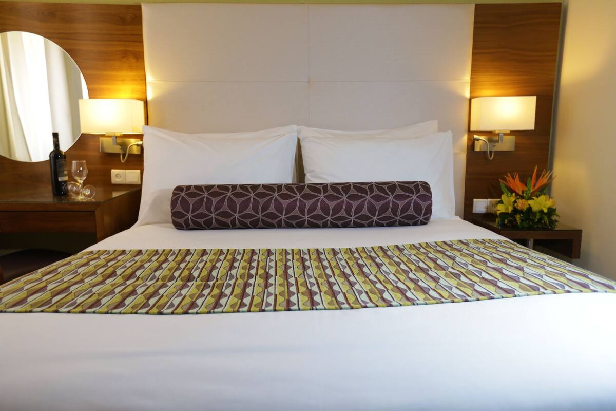Habitació d'hotel amb un llit gran, dues làmpades de nit, una tauleta de nit amb una ampolla i gots, i un mirall.