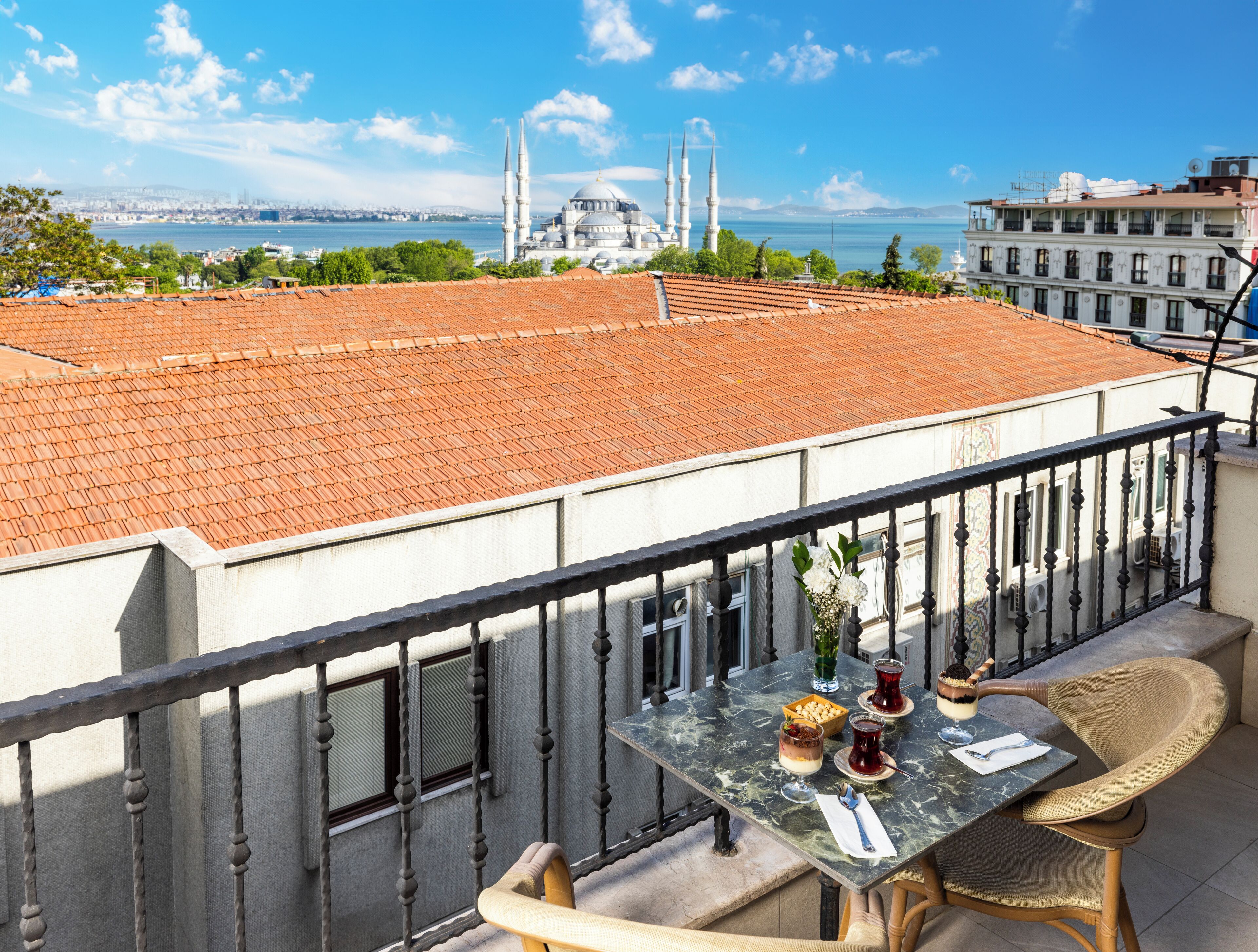 Un balcón con una mesa lista para comer, con vista a un paisaje urbano que incluye una mezquita destacada y una masa de agua.