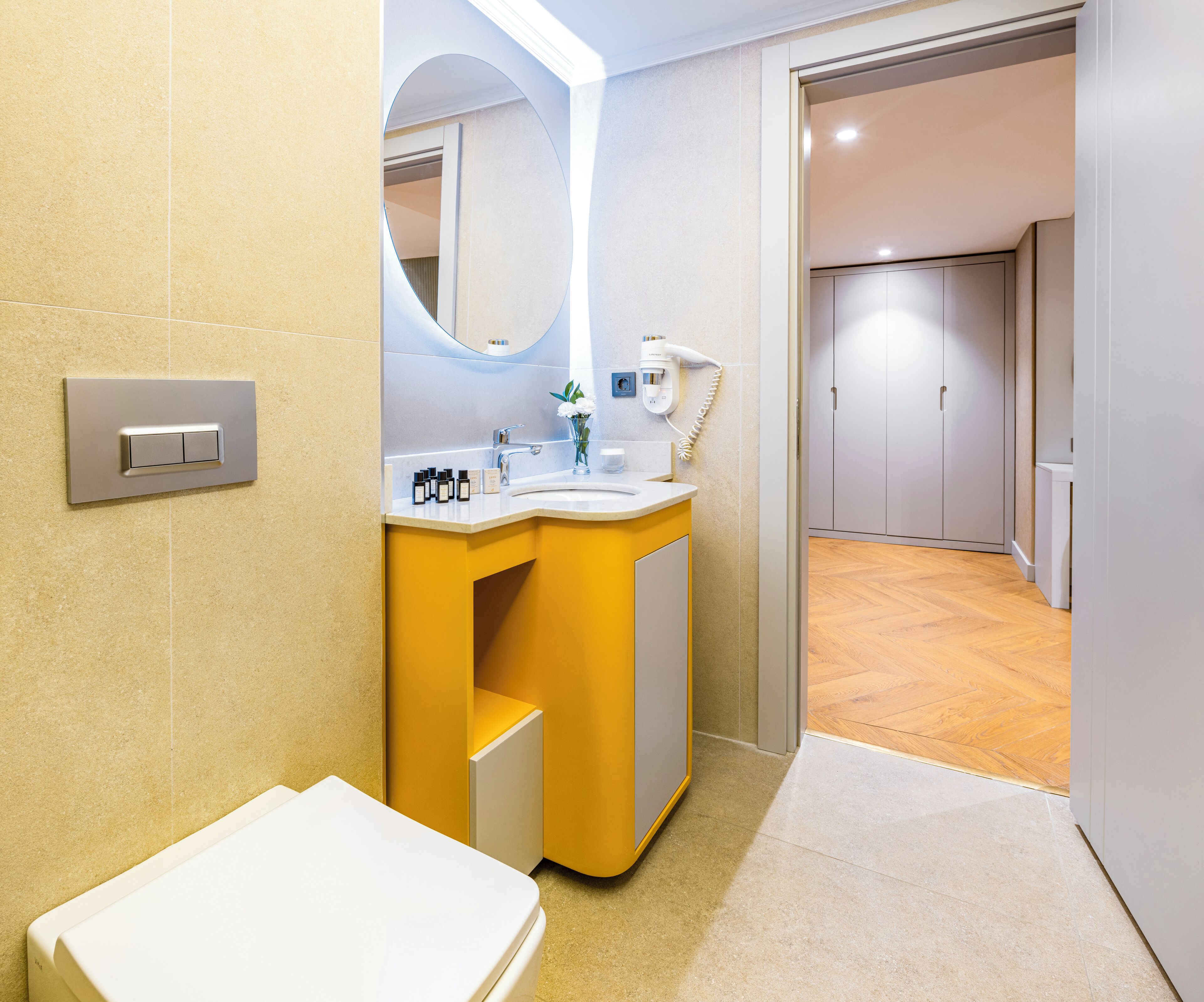 Ein modernes Badezimmer mit einem gelben Waschtisch, einem weißen Waschbecken und einer Toilette. Darüber hängt ein Spiegel mit runder Fassung, und eine weiße Handtuch hängt an der Wand.