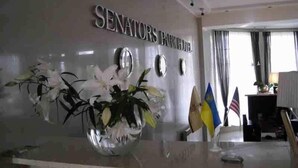 Un comptoir d'accueil avec une pancarte indiquant « HÔTEL SENATORS PARK ». On y voit des drapeaux et un vase rempli de fleurs.