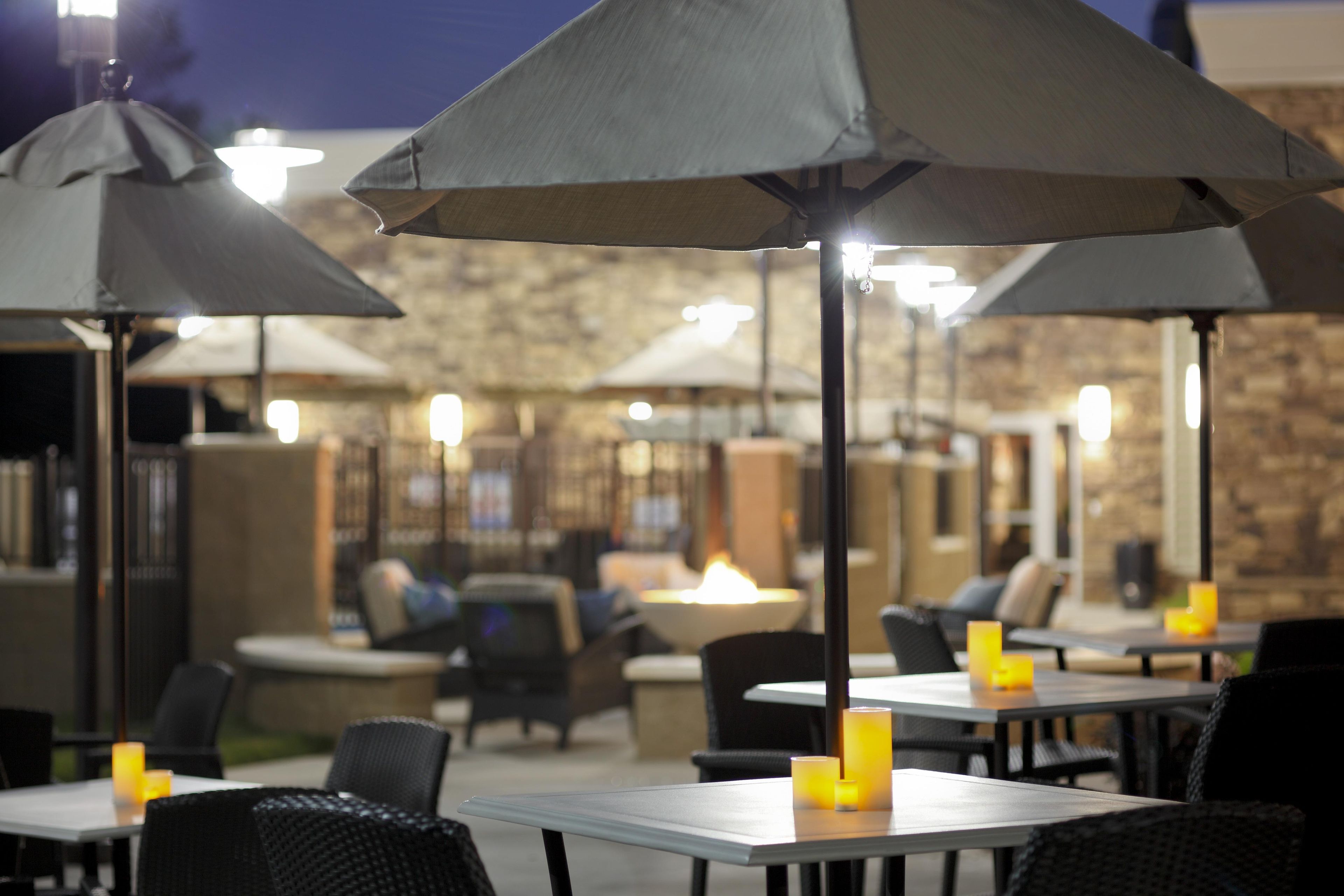 An outdoor seating area with tables and umbrellas, a stone wall in the background, and a fire feature.